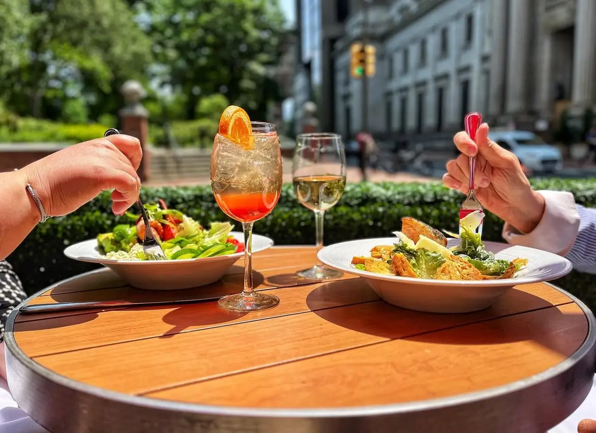 Two people enjoying fresh salads and spritzes at an outdoor bistro table on a sunny day. A sparkling orange cocktail and glass of white wine add to the relaxed city lunch vibe.