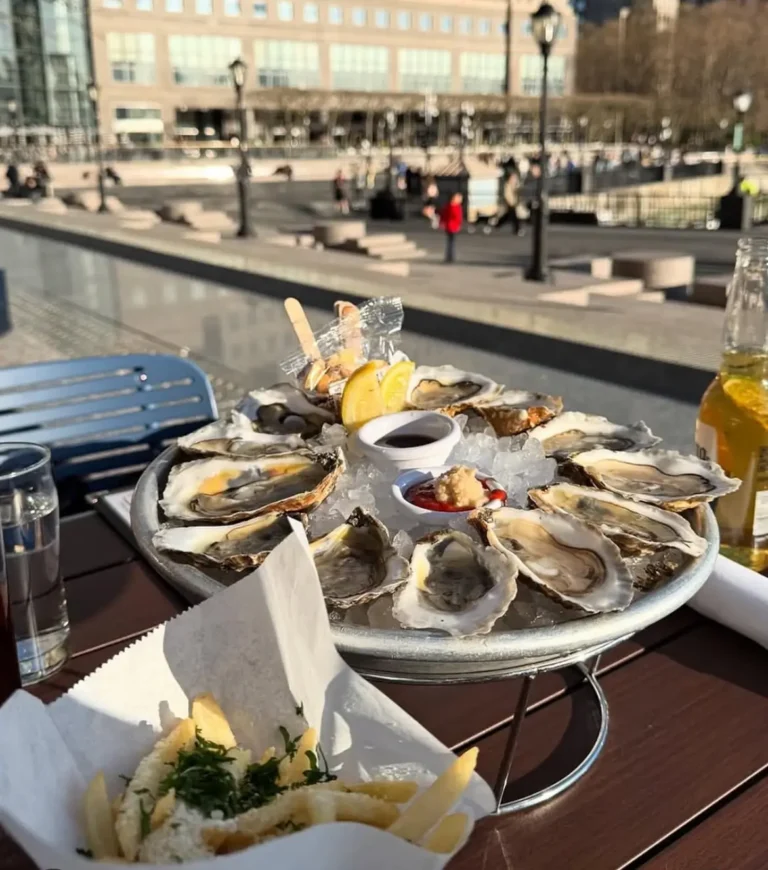A large platter of oysters on ice, served with lemon wedges, cocktail sauce, and mignonette, set on an outdoor table with a view of a sunny plaza. A side of truffle fries and bottles of beer complete the casual seafood spread.