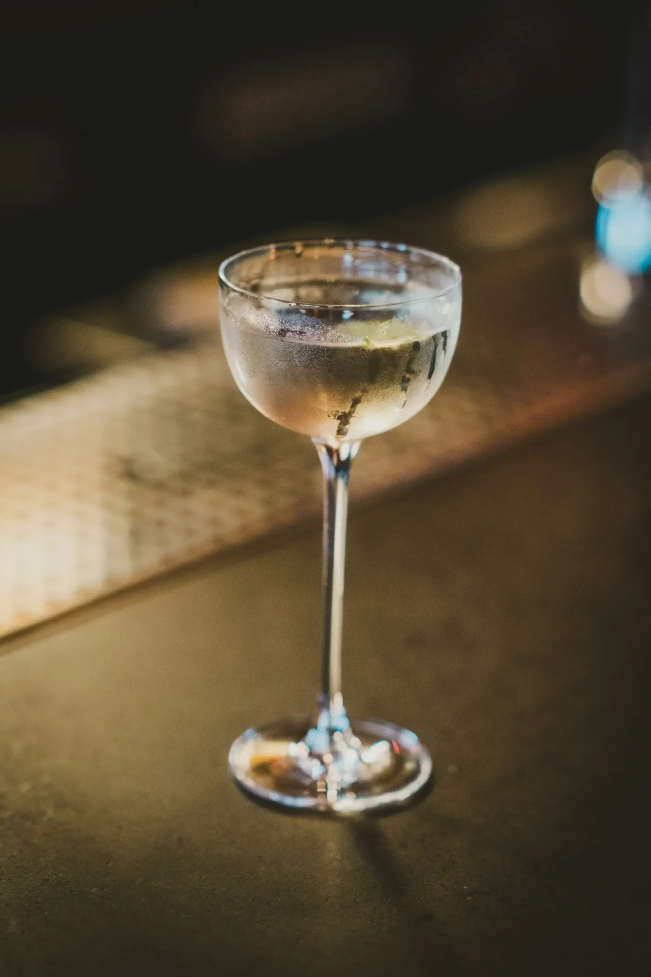 A chilled coupe glass filled with a clear cocktail, condensation dripping down the side, resting on a dark bar counter in a dimly lit setting.