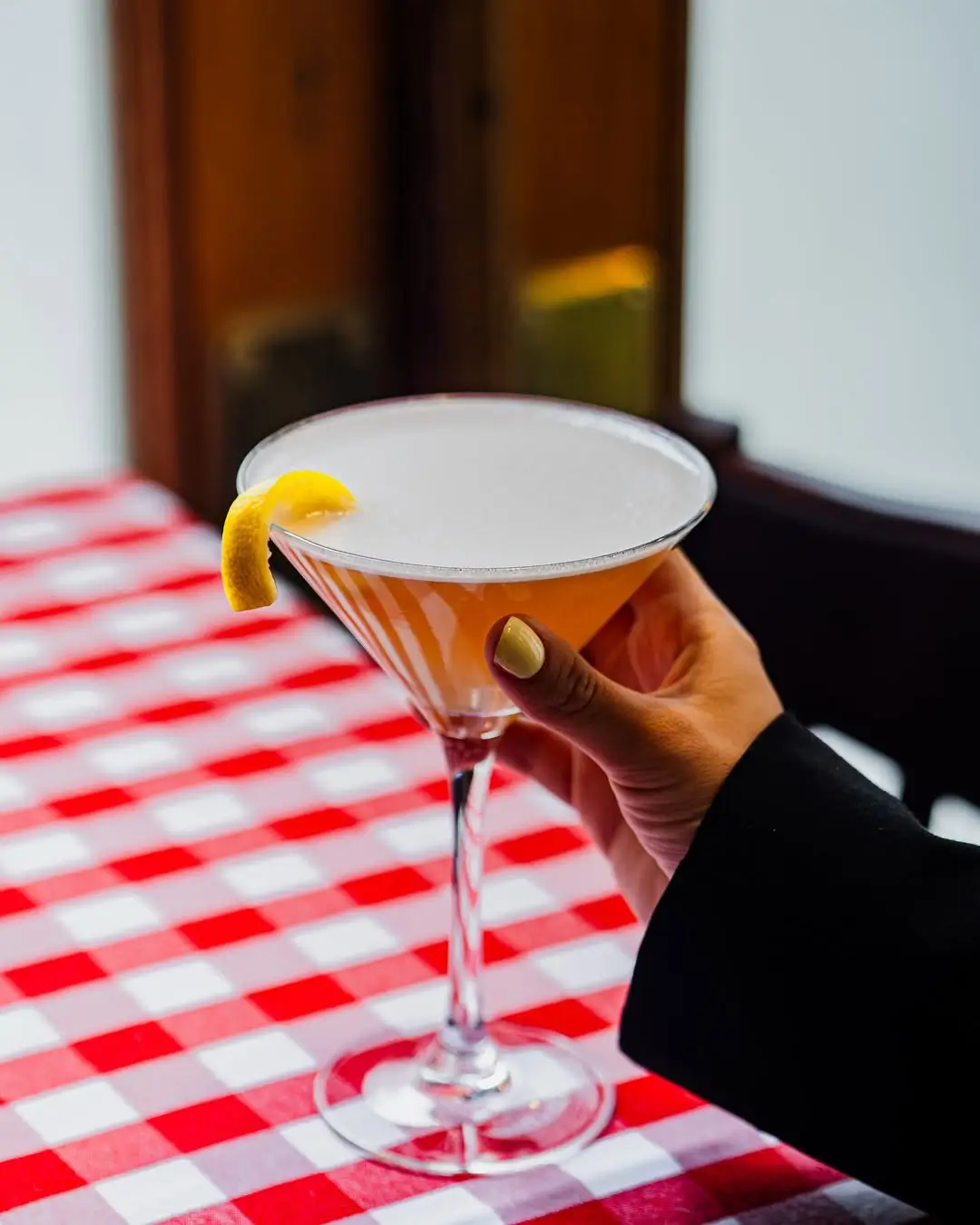 A hand holding a frothy cocktail in a martini glass garnished with a lemon twist, set against a classic red-and-white checkered tablecloth.