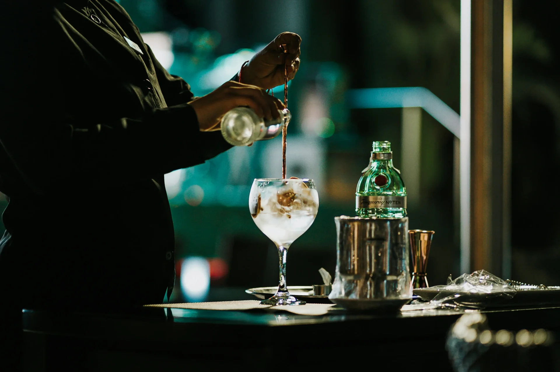 A bartender pours tonic into a large stemmed glass filled with ice and botanicals, crafting a cocktail under moody bar lighting. A green San Pellegrino bottle and bar tools sit nearby.